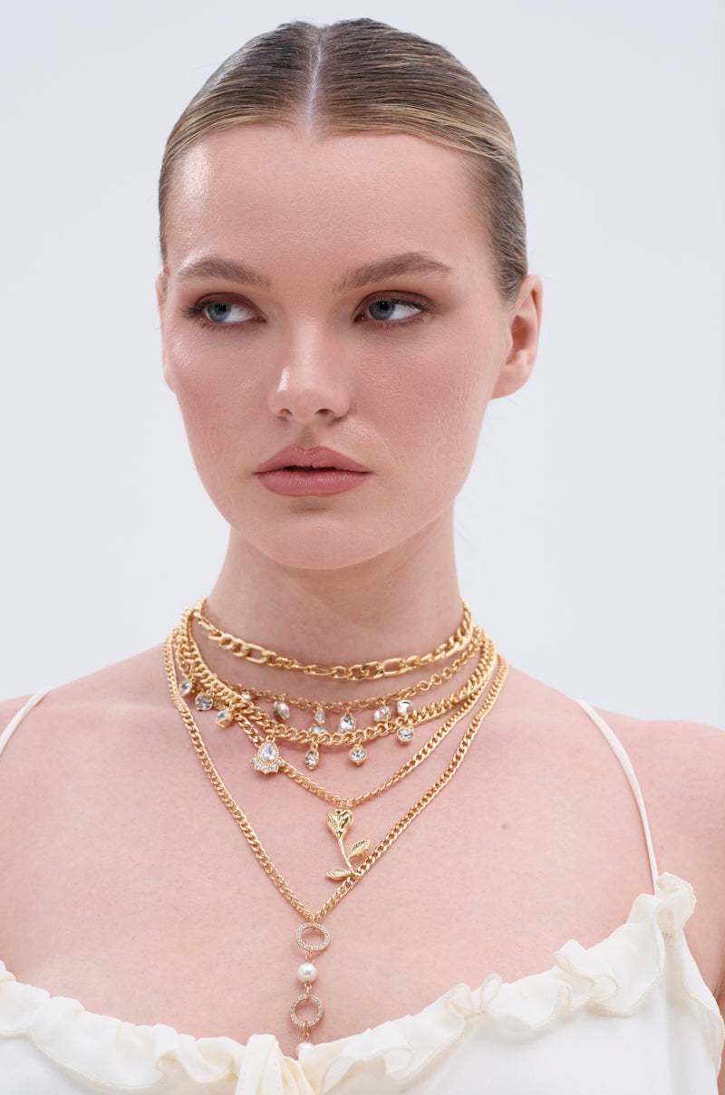 A woman with slicked-back blonde hair wears the GOING ON A TRIP GOLD NECKLACE SET—multi-layered chains with crystals and a rose charm—paired with a white ruffled strappy top as she gazes to the side against a light background. 0