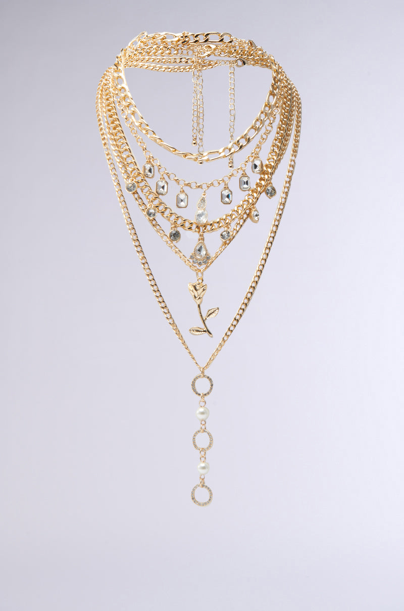 The GOING ON A TRIP GOLD NECKLACE SET, featuring multi-layered gold chains, crystal-like pendants, and a rose charm, is elegantly displayed against a plain light gray background. 1