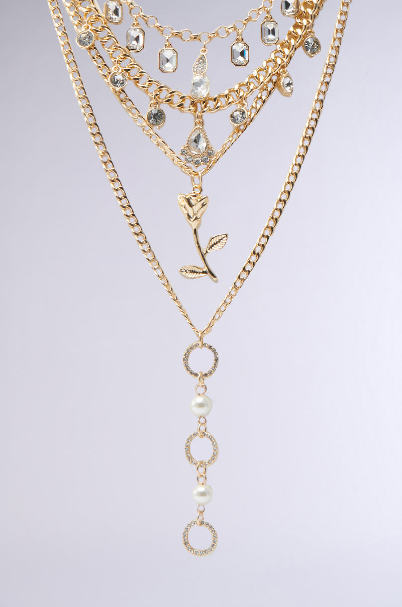 The GOING ON A TRIP GOLD NECKLACE SET features layered gold chains with crystals, gems, a central rose charm, pearl drop chain, and textured gold rings, all beautifully displayed on a light grey background. 2