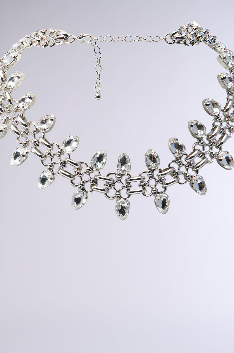 The Real Will Know Necklace is a faux silver piece featuring interlocking hardware with two layers of chains, adorned with teardrop-shaped clear gemstones, set against a light gray background. 2