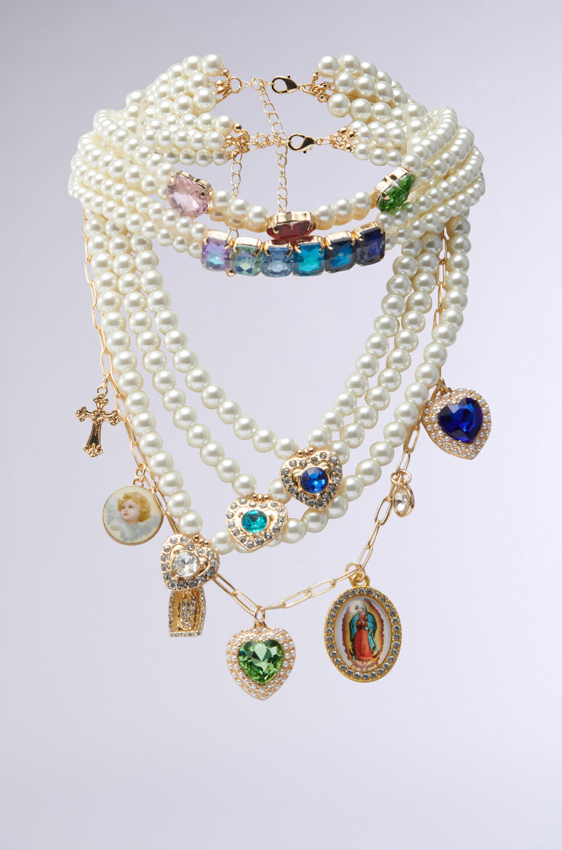 The FEEL MY VIBE NECKLACE is a multi-layered piece with strands of pearls, gold chains, and gemstone pendants like religious icons, heart-shaped gems, and a cross. Each strand showcases unique ornate charms and jewels.