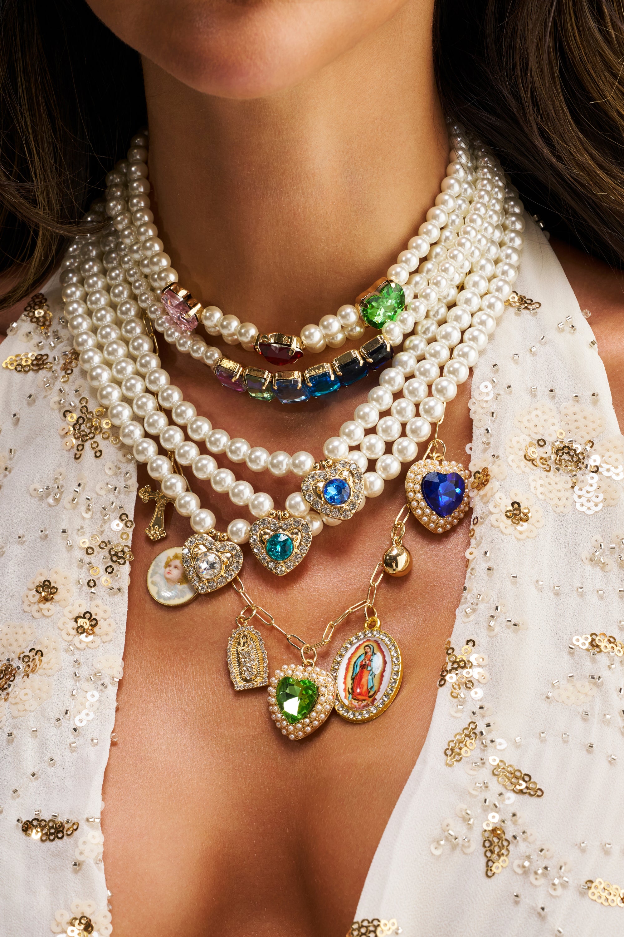 A woman wears the FEEL MY VIBE NECKLACE, a layered pearl piece featuring colorful gemstone pendants and religious charms, styled with a white sequin-embellished top.