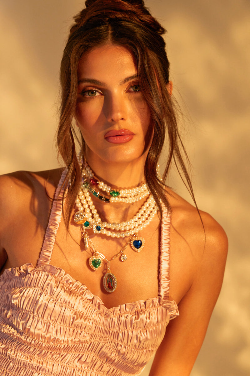 A woman with long brown hair in an elegant updo wears a pink ruched dress and the FEEL MY VIBE NECKLACE, showcasing pearls, gemstone pendants, and colorful jewels. The soft lighting enhances the warm and sophisticated ambiance.