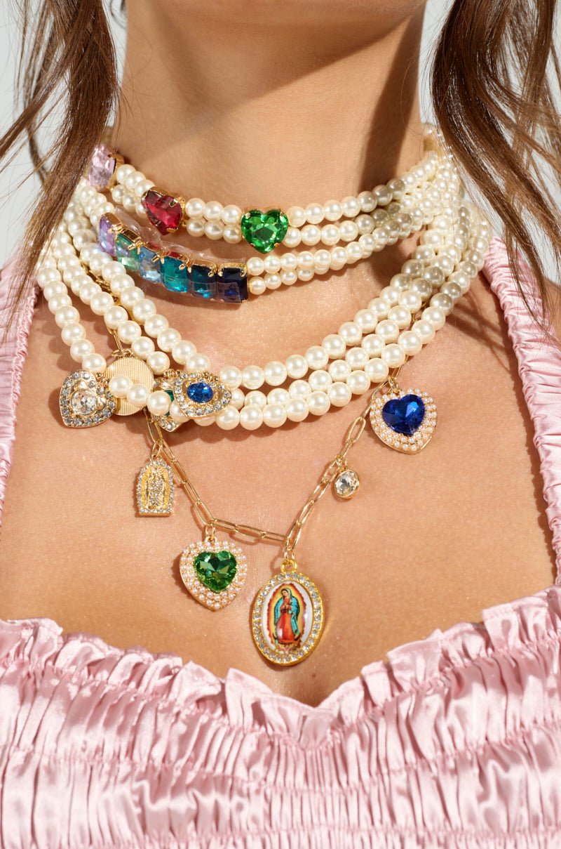 Someone wears the FEEL MY VIBE NECKLACE, a choker of layered pearls with colorful heart-shaped gems and small religious pendants. They pair it with a pink ruched top. 3