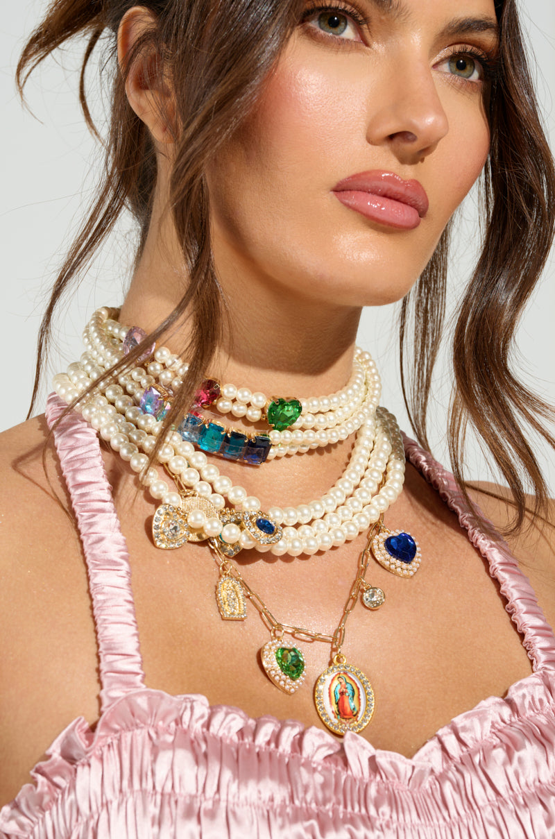 A woman wears the FEEL MY VIBE NECKLACE, layered with pearl strands and colorful gemstone pendants alongside religious charms, enhancing her wavy brown hair. She pairs it with a pink ruched satin top against a neutral backdrop, radiating elegance.