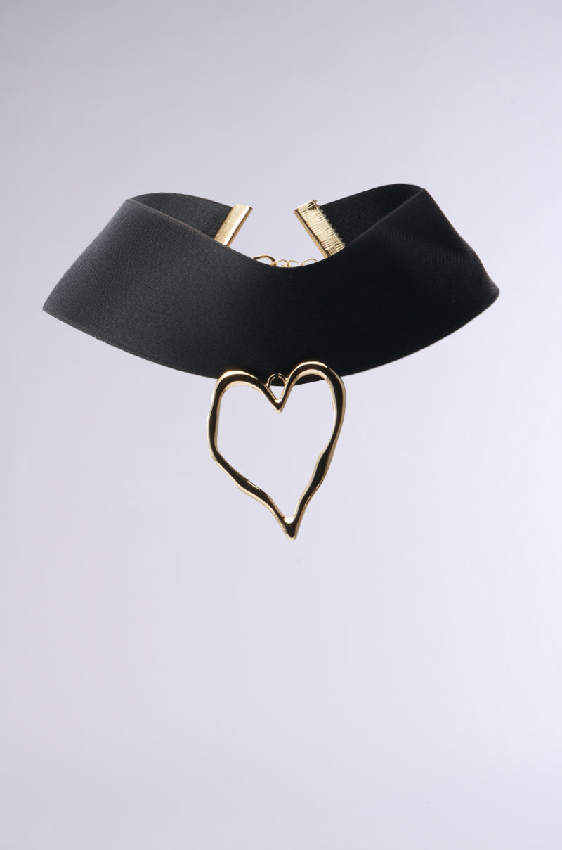 The FEEL THE LOVE CHOKER NECKLACE features a satin black ribbon with a gold-toned heart pendant at its center, showcasing an uneven, organic shape against a plain, light gray background.