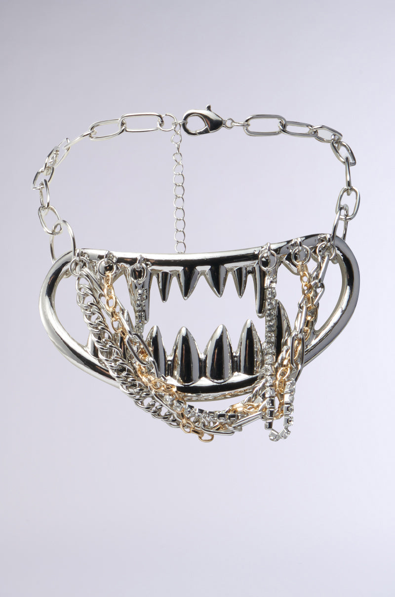 The BITE ME NECKLACE is a bold piece with large metallic spikes in a zigzag pattern, interspersed with faux silver chains and rhinestones, mimicking jagged teeth on a linked chain.
