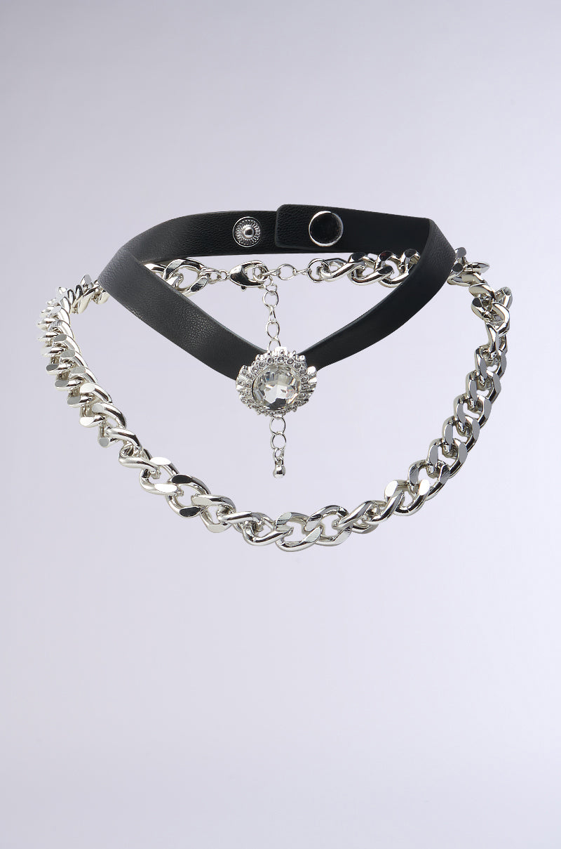 The IN THE SHOW CHOKER NECKLACE SET features a wide black faux leather strap with a metal button clasp, a large Cuban chain, and a decorative charm with dangling silver-toned ornament, all set against a plain light gray background. 1