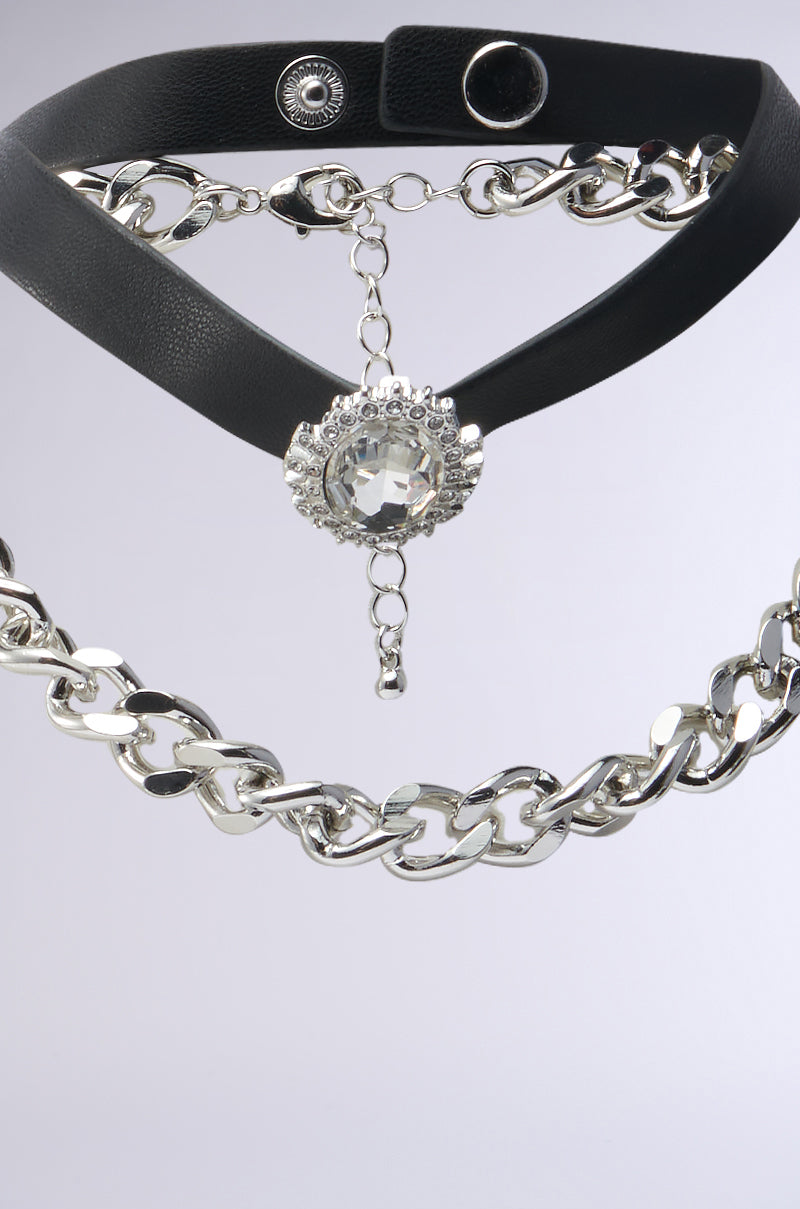 The IN THE SHOW CHOKER NECKLACE SET features a black faux leather choker with a large circular rhinestone centerpiece and an interlocking design silver chain. They are elegantly displayed on a simple gray background for a chic style. 3