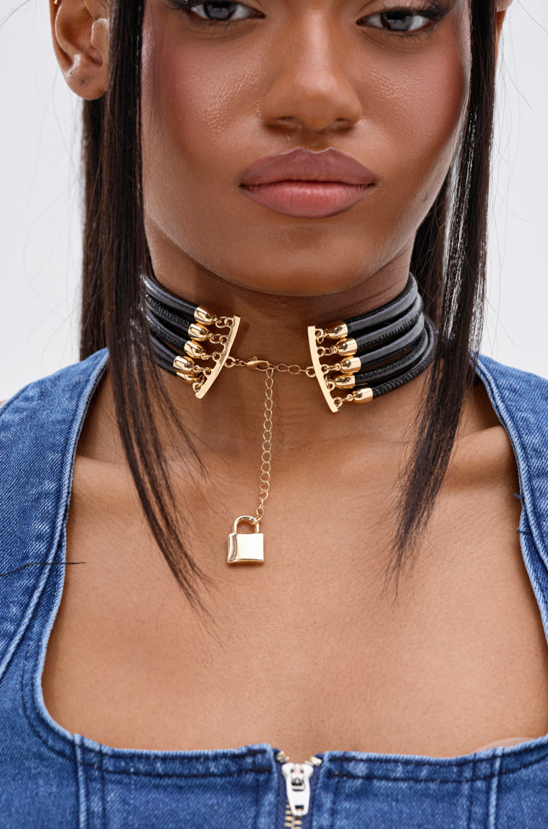 A woman wears the KEY TO MY HEART NECKLACE, a multi-strand choker with gold-tone hardware and a gold padlock pendant, paired with a blue denim zip-up top.