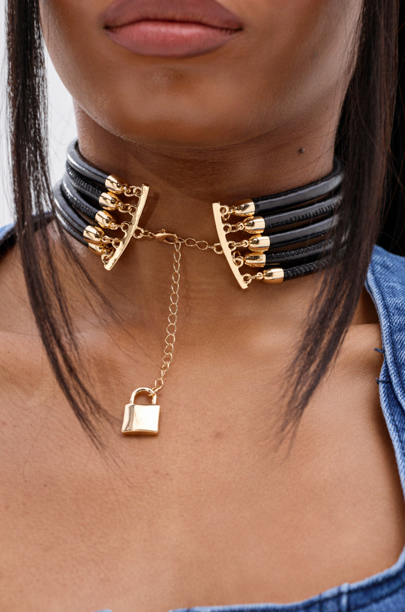 A close-up of a person with dark hair and a blue garment wearing the KEY TO MY HEART NECKLACE, featuring multiple strands with gold-tone hardware, decorative metal tips, a central gold chain, and a small padlock pendant.