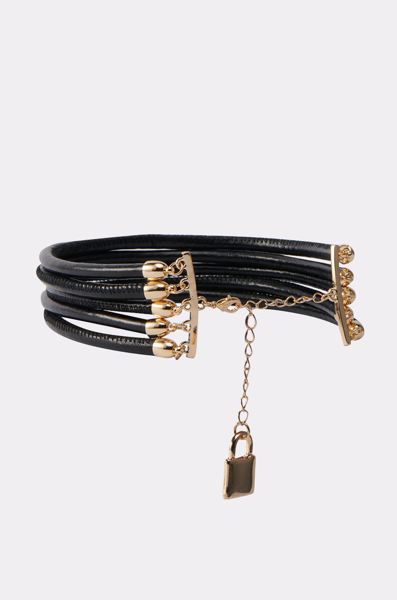 The KEY TO MY HEART NECKLACE features black multi-strand leather, gold-tone end pieces, dangling gold beads, an adjustable chain, and a small gold padlock charm. Displayed on a plain light grey background.