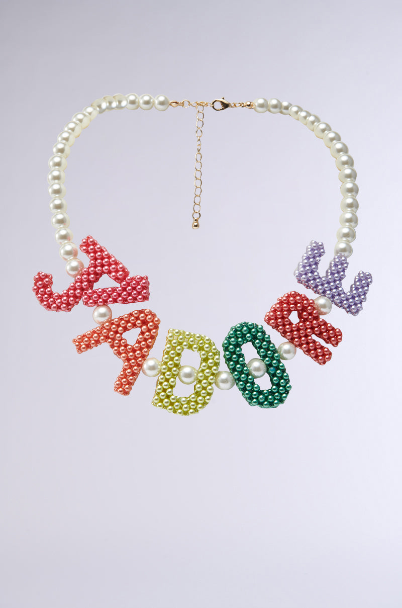 The JADORE NECKLACE showcases delicate pearls and vibrant beaded letters spelling "JADORE," displayed on a light gray background and finished with an adjustable lobster clasp for a customized fit.