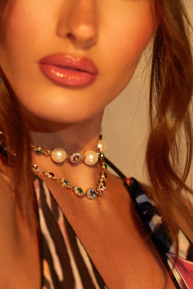 A woman wears a striped top and the CANDY CRUSHIN' NECKLACE SET with multicolor jewels. Her glossed lips complement her wavy brown hair that cascades over her shoulders, partially framing her face.