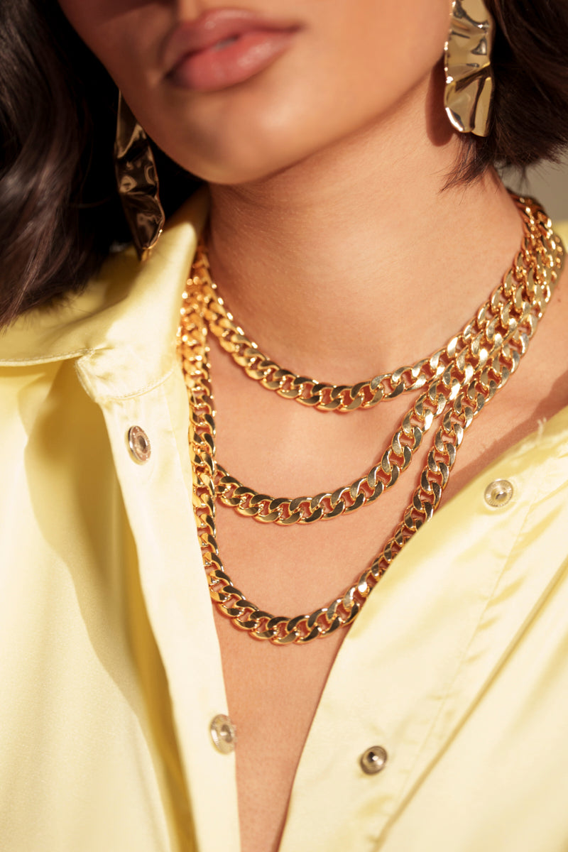 A woman, shown from the lips down, wears layered gold chains, including the "BUY ME THIS NECKLACE" with a padlock charm, styled with textured gold earrings and an unbuttoned pale yellow satin shirt.