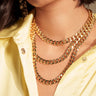 A woman, shown from the lips down, wears layered gold chains, including the "BUY ME THIS NECKLACE" with a padlock charm, styled with textured gold earrings and an unbuttoned pale yellow satin shirt.