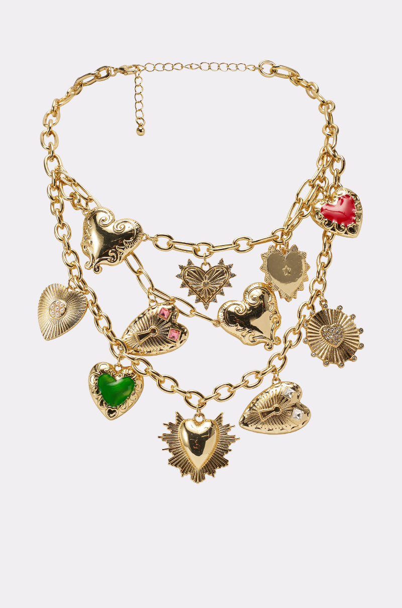 The TAKE ME TO MILAN NECKLACE is a layered gold necklace with multiple heart-shaped and ornate charms, featuring green and red enamel details on chunky chain tiers, shown against a white background.