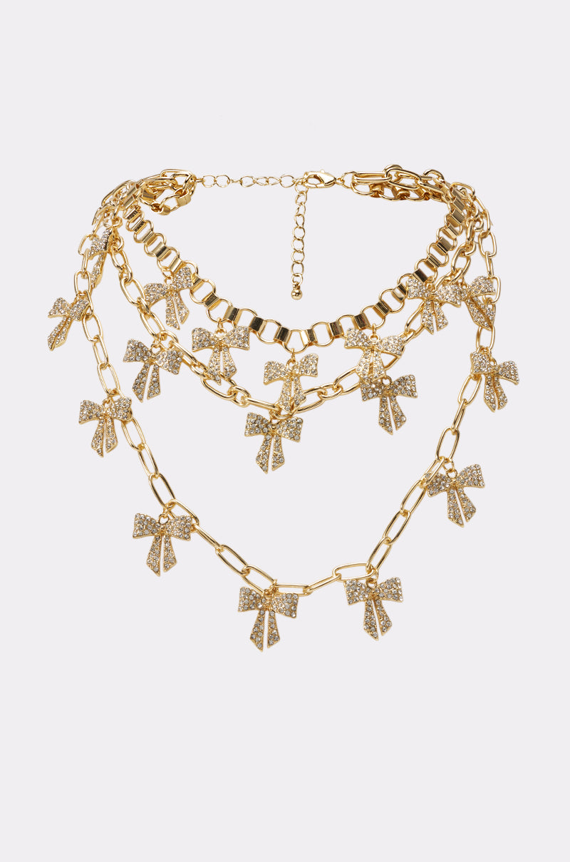 Introducing the TIED IN A BOW NECKLACE: a layered gold necklace showcasing three delicate chains, each embellished with sparkling bow-shaped pendants encrusted with rhinestones. This rhinestone bow necklace features an adjustable clasp for the perfect fit and shines elegantly against any background—ideal as festival jewelry or for adding a glamorous touch to every occasion.