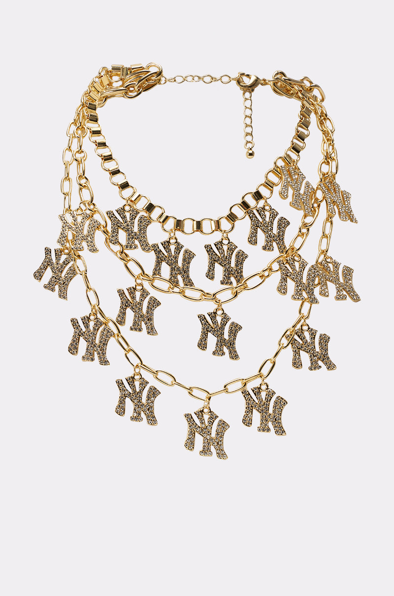 The IN THE CITY LAYERED NECKLACE features gold chains with sparkling "NY" rhinestone letter charms, creating a stylish look that stands out beautifully against a plain white background. 1