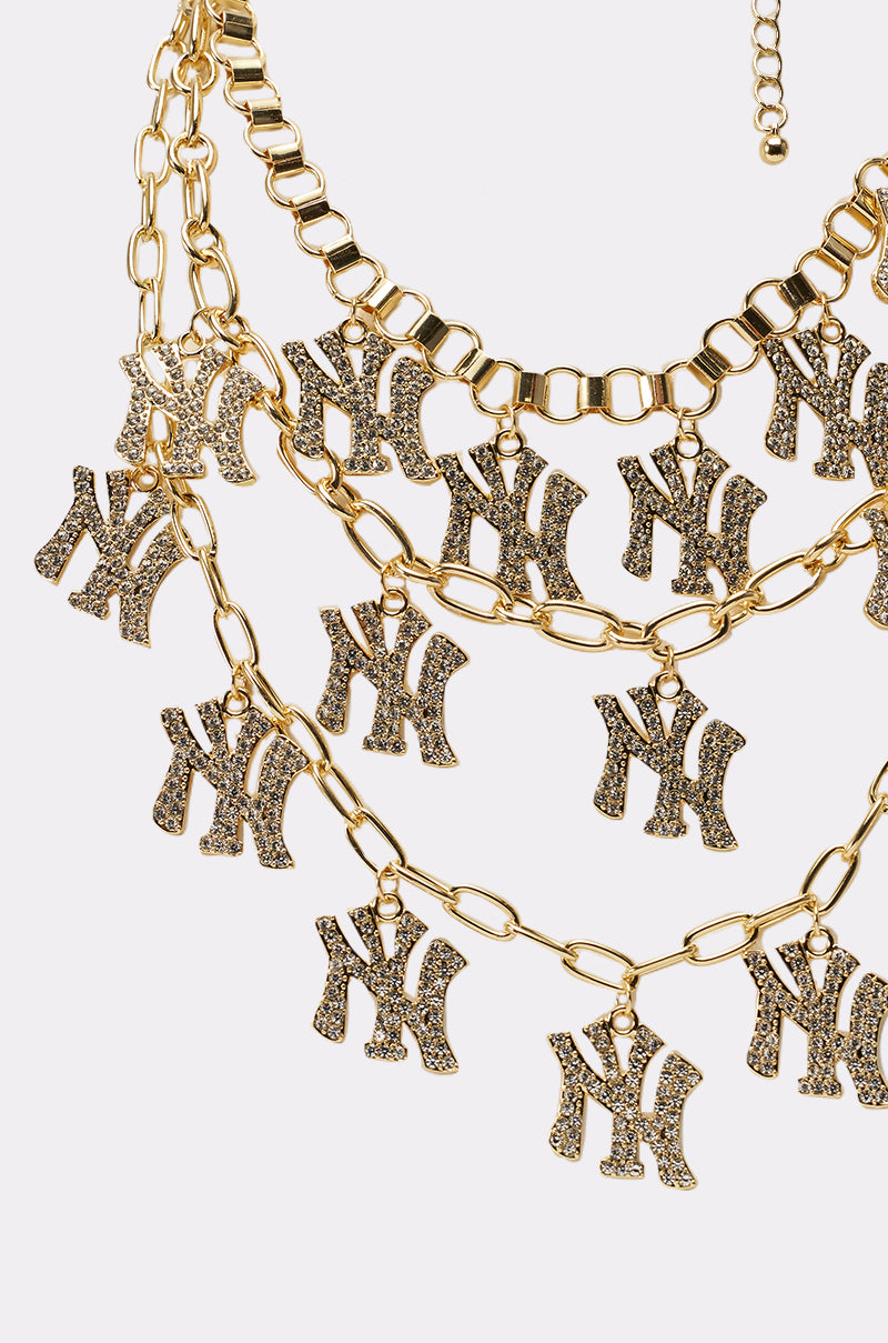 The IN THE CITY LAYERED NECKLACE features gold chains with rhinestone "NY" letter charms on each tier, displayed against a plain white background. 2