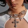 A woman wears a sheer white top and the FOREVER PROTECTED NECKLACE—a bold silver-tone piece with three rhinestone cross pendants. Her striking makeup and serious expression highlight her standout statement jewelry.