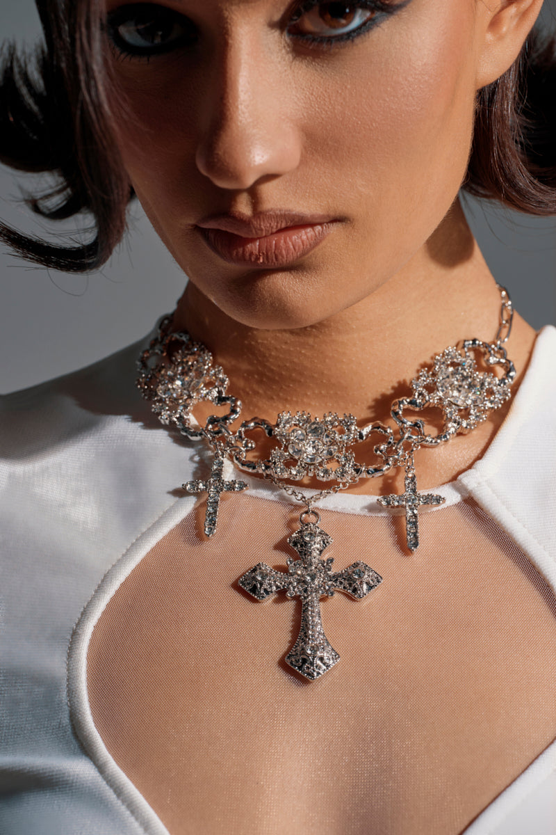 A woman wears a sheer white top and the FOREVER PROTECTED NECKLACE—a bold silver-tone piece with three rhinestone cross pendants. Her striking makeup and serious expression highlight her standout statement jewelry. 1