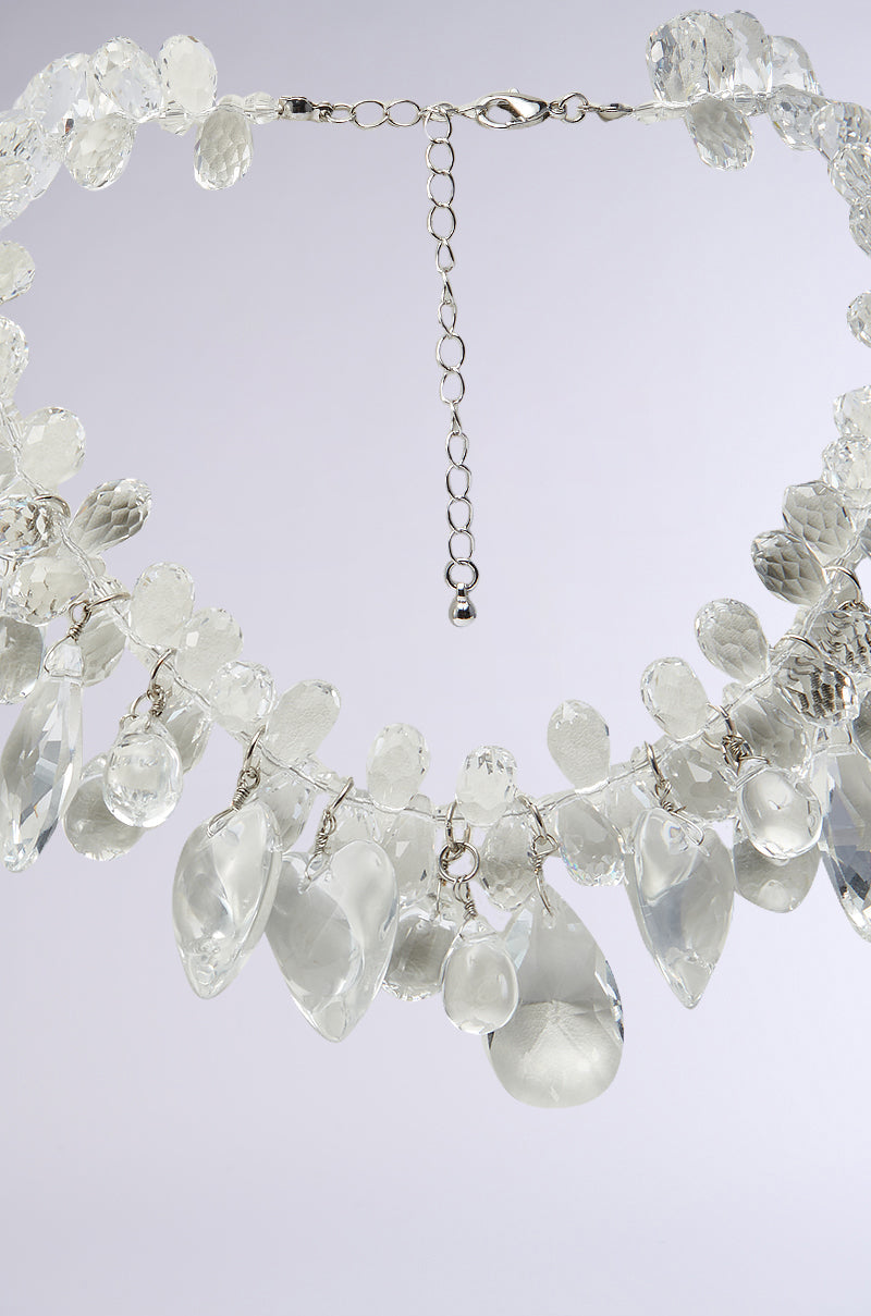 A close-up of the CLEAR TO ME NECKLACE, featuring faceted and smooth clear heart-shaped drops on a silver chain with an adjustable clasp, set against a light gray background. 2