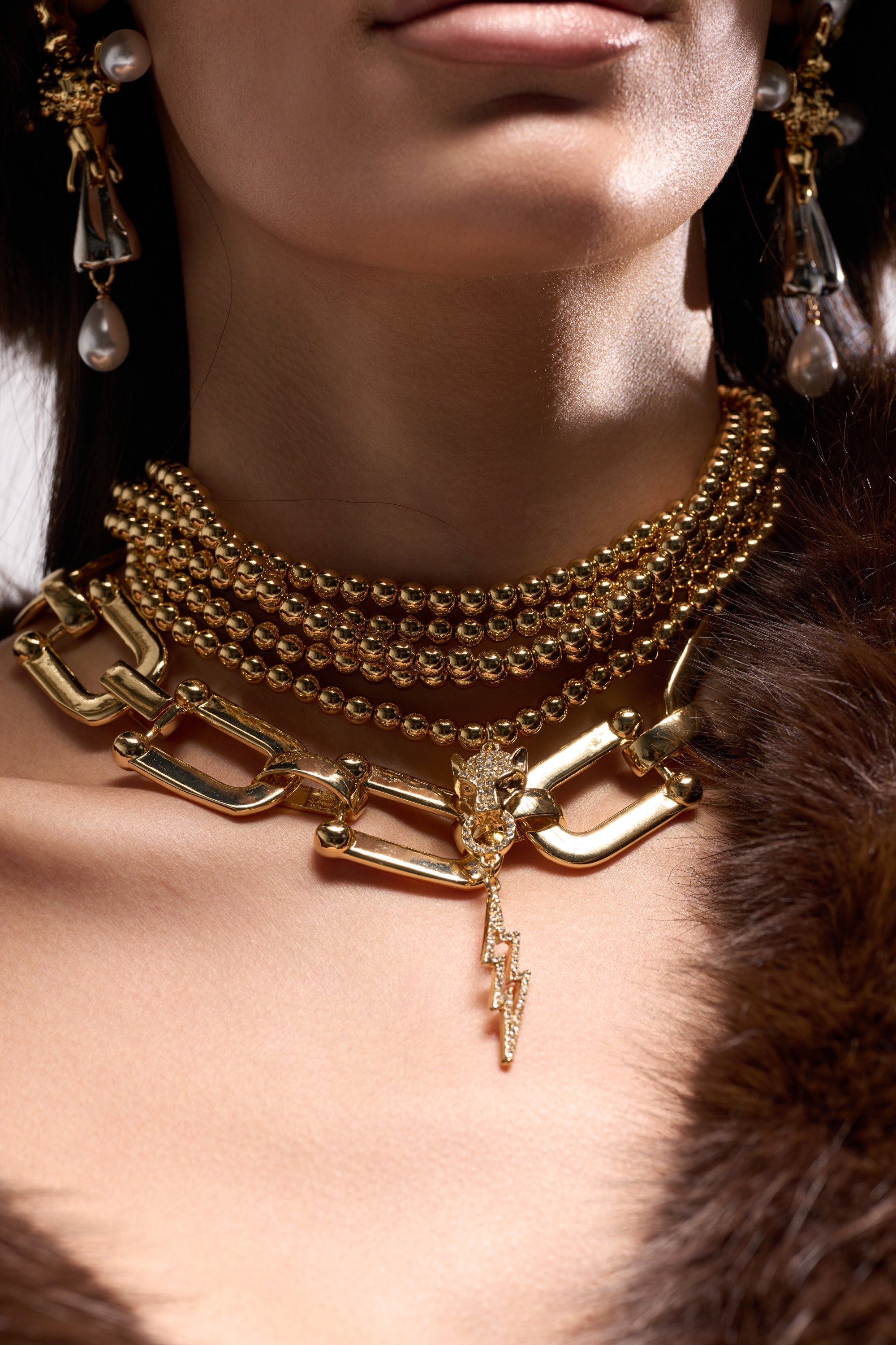 A woman wears the GOLD QUEEN NECKLACE SET featuring layered gold chains, a panther head pendant, a lightning bolt pendant, and pearl drop earrings, styled with a brown fur garment; only her lower face and neck are visible. 0