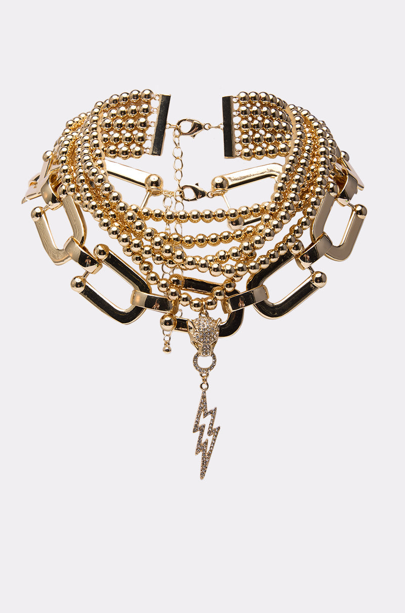 The GOLD QUEEN NECKLACE SET is a bold, layered gold choker featuring chunky chains, round beads, rectangular links, and a rhinestone lightning bolt charm, shown against a plain white background. 2