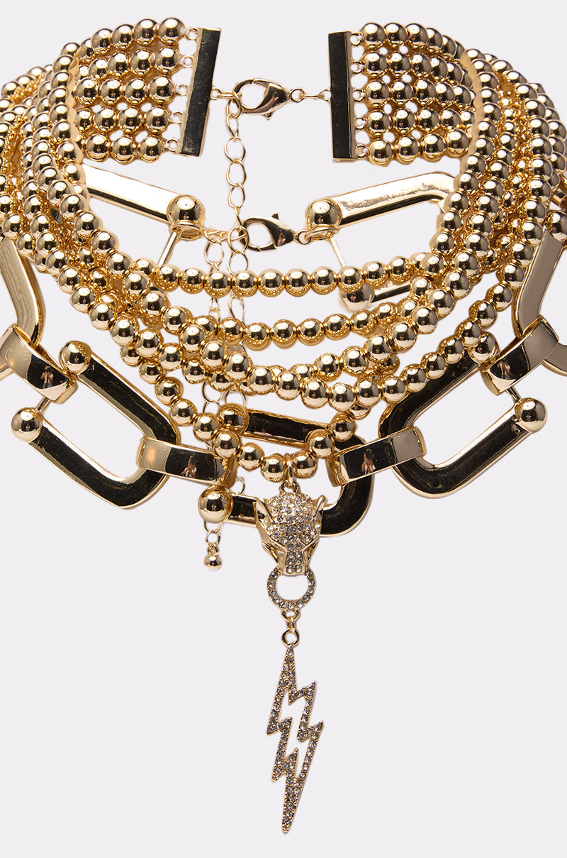 The GOLD QUEEN NECKLACE SET is a layered gold choker with gold-tone chains, assorted beads, geometric links, and a rhinestone-studded dangling lightning bolt charm. 3