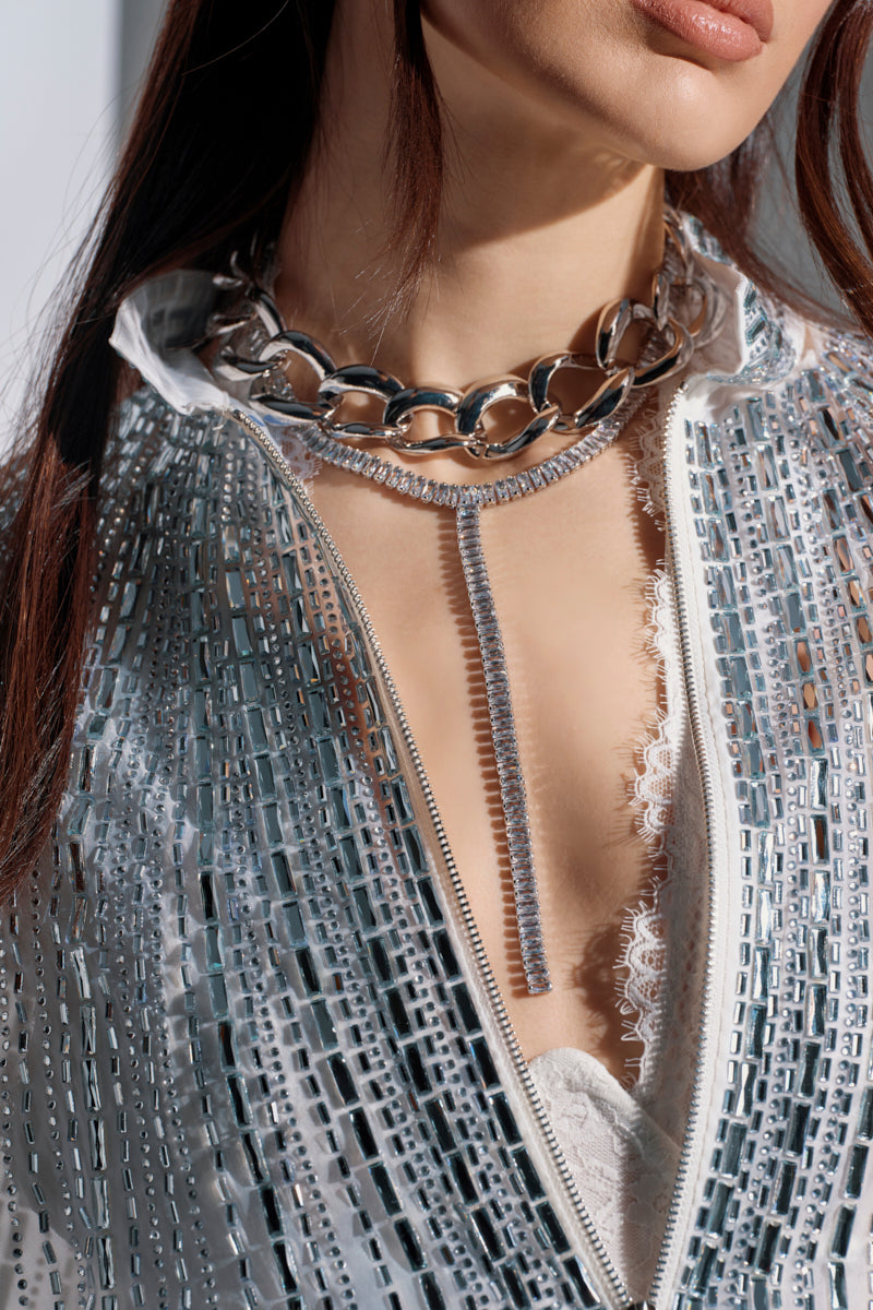 A woman wears a mirrored jacket, unzipped to show a white lace top. She accessorizes with the PAIR ME NECKLACE SET: a chunky silver-tone chain and a rhinestone Y-drop strand with adjustable closures.