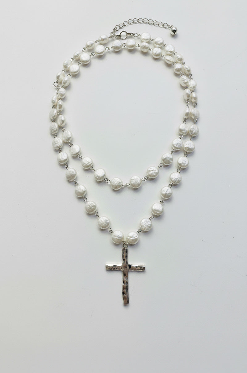 The HOLY MOTHER OF PEARLS NECKLACE features two layers of round white pearls, accented by a striking cross pendant on the longer strand, elegantly displayed against a plain white background.