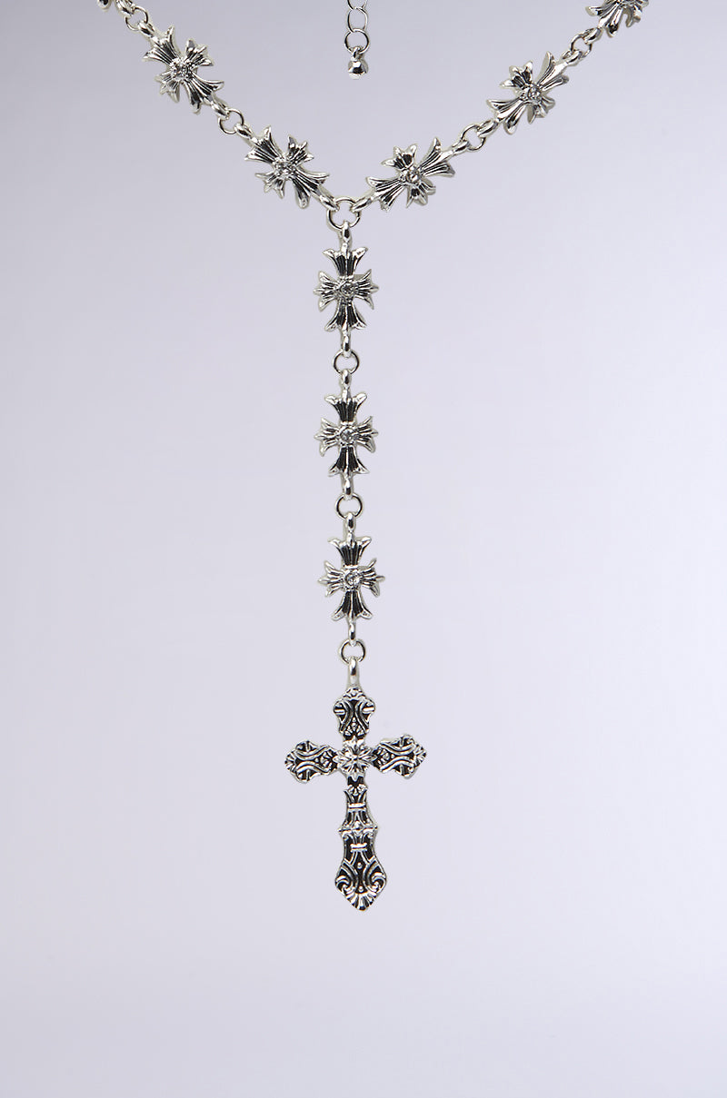 The DEDE CROSS NECKLACE is a silver rosary-style piece with ornate floral beads accented by rhinestone centers and a decorative cross pendant, shown against a plain light background.