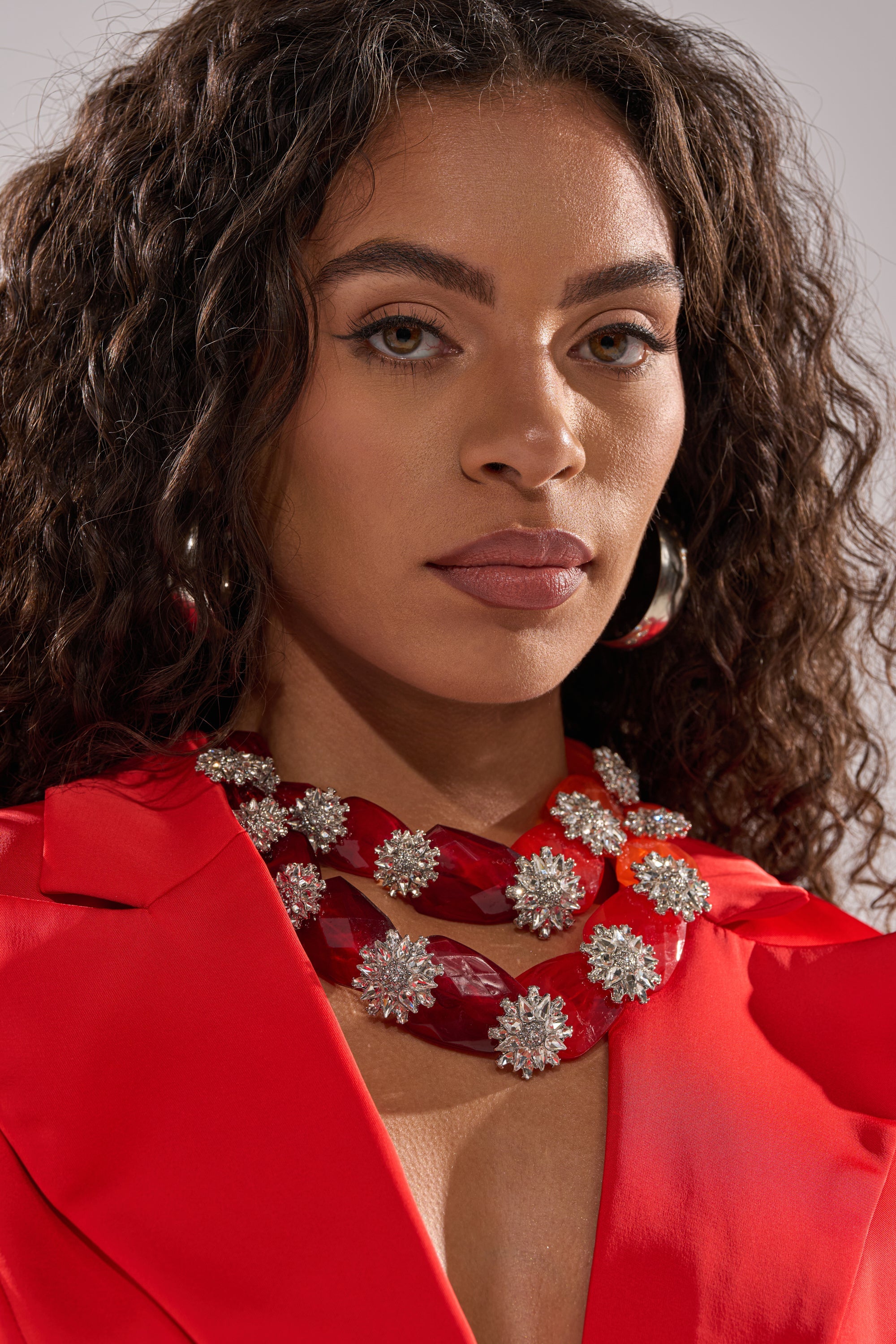 A woman with curly hair wears a bright red outfit, large silver hoops, and the ESTABLISHED NECKLACE featuring sparkling silver flower-shaped ornaments. She looks confidently at the camera. 1