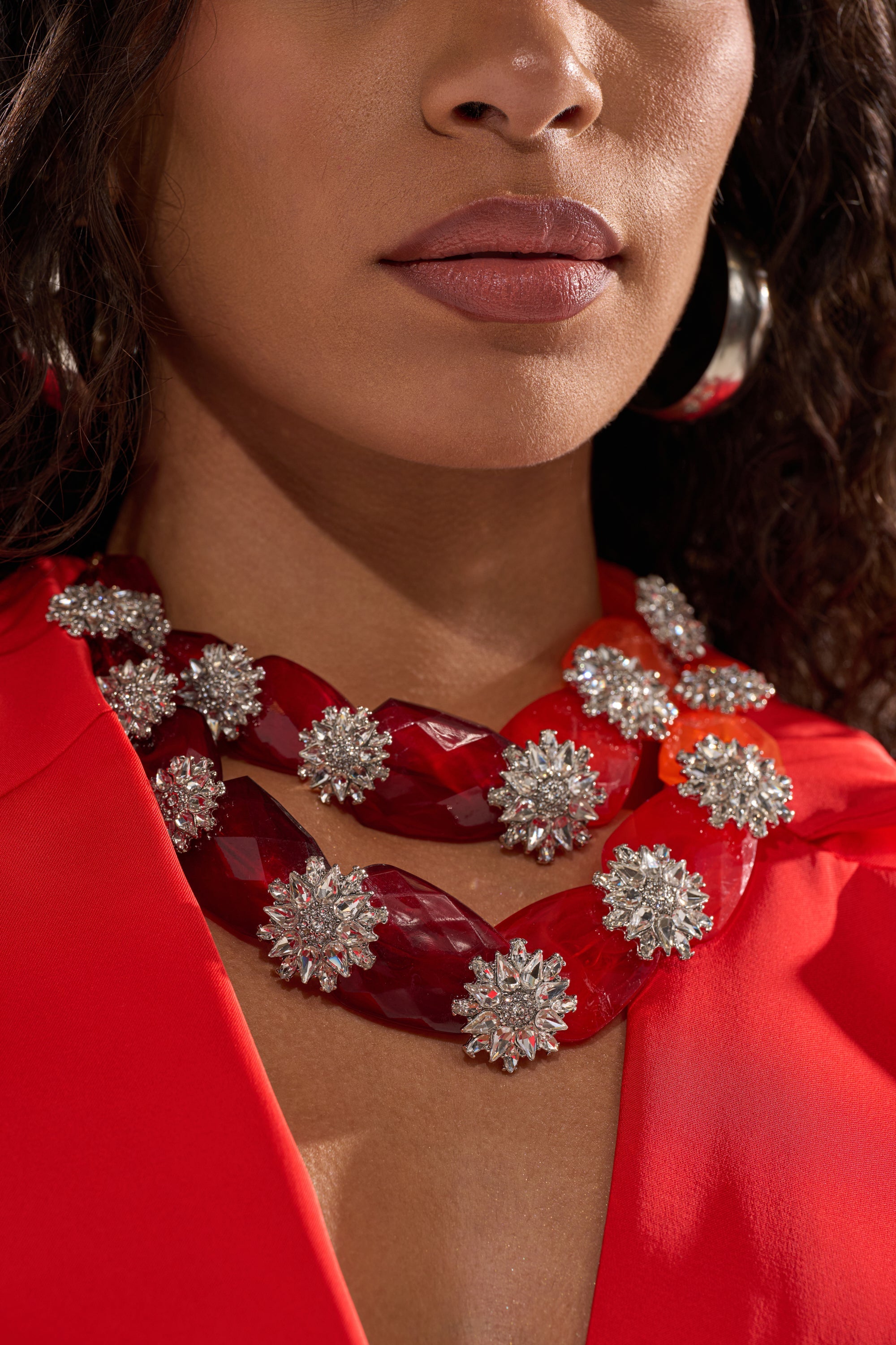 A woman with curly dark hair wears a bright red top and the ESTABLISHED NECKLACE, a layered red piece with silver floral details, along with large silver hoop earrings. The image is cropped at her nose and shoulders. 2