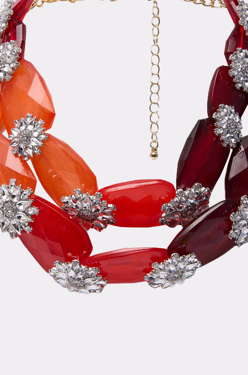 Close-up of the ESTABLISHED NECKLACE: a chunky statement piece with large, faceted red and orange beads separated by sparkling silver floral accents. Necklace features a gold chain extender, shown on a plain white background. 3