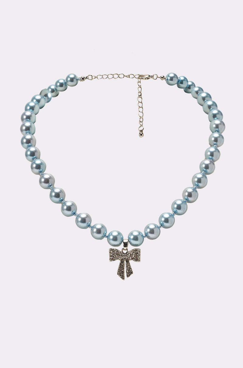 The KRISTEN NECKLACE showcases large, glossy light blue pearls with a sparkling rhinestone bow charm as its gold pendant, set against a white background. 1