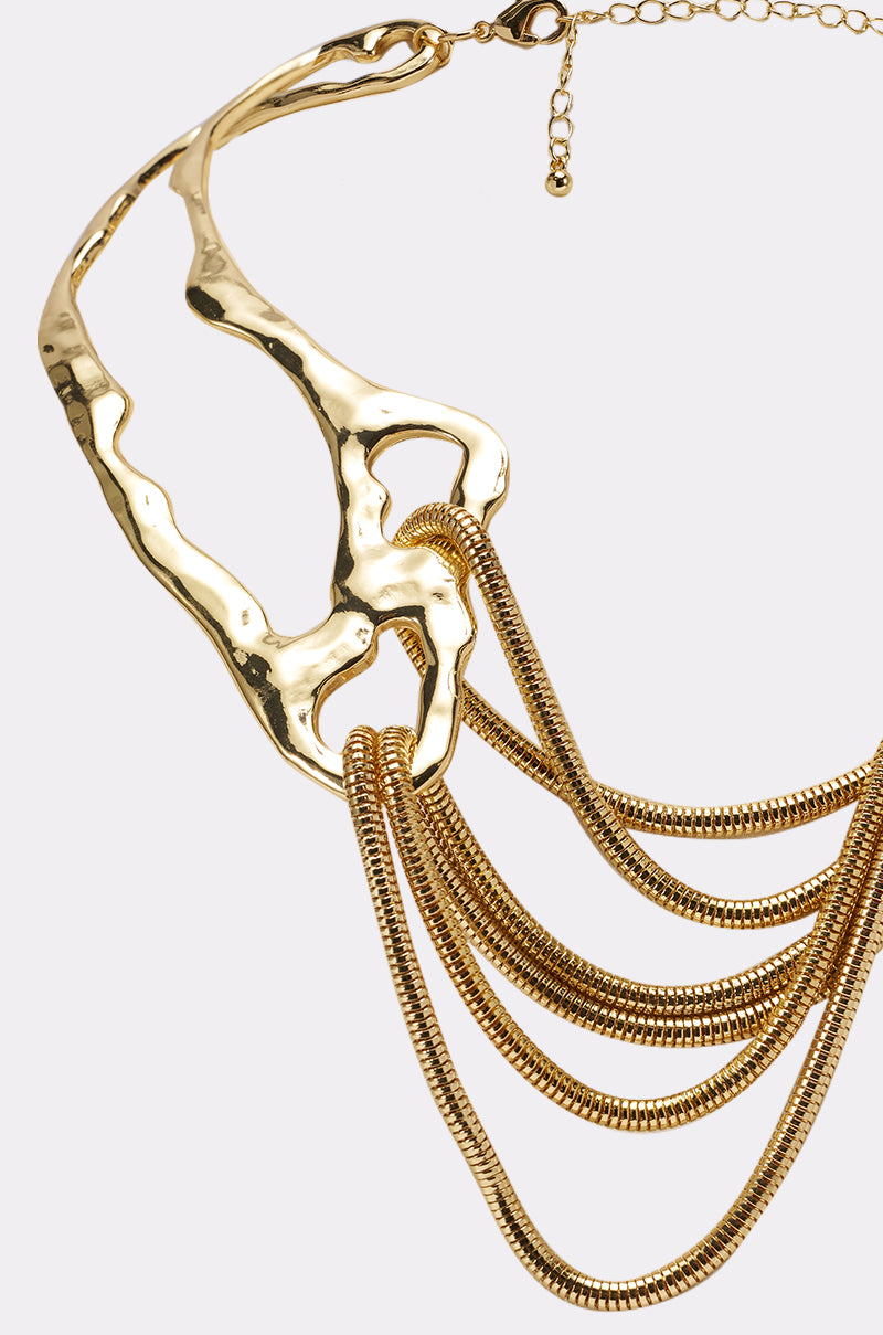 Close-up of the SO DECO NECKLACE, a sculptural gold statement piece featuring an abstract metal plate with several draped, textured gold chains against a plain background. 3