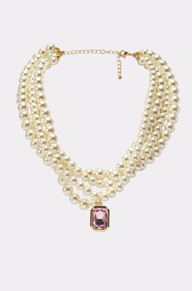 The MARIE LAYERED NECKLACE features three strands of ivory faux pearls, a bold pink gemstone pendant at the center, and is finished with a gold clasp and extender. 1