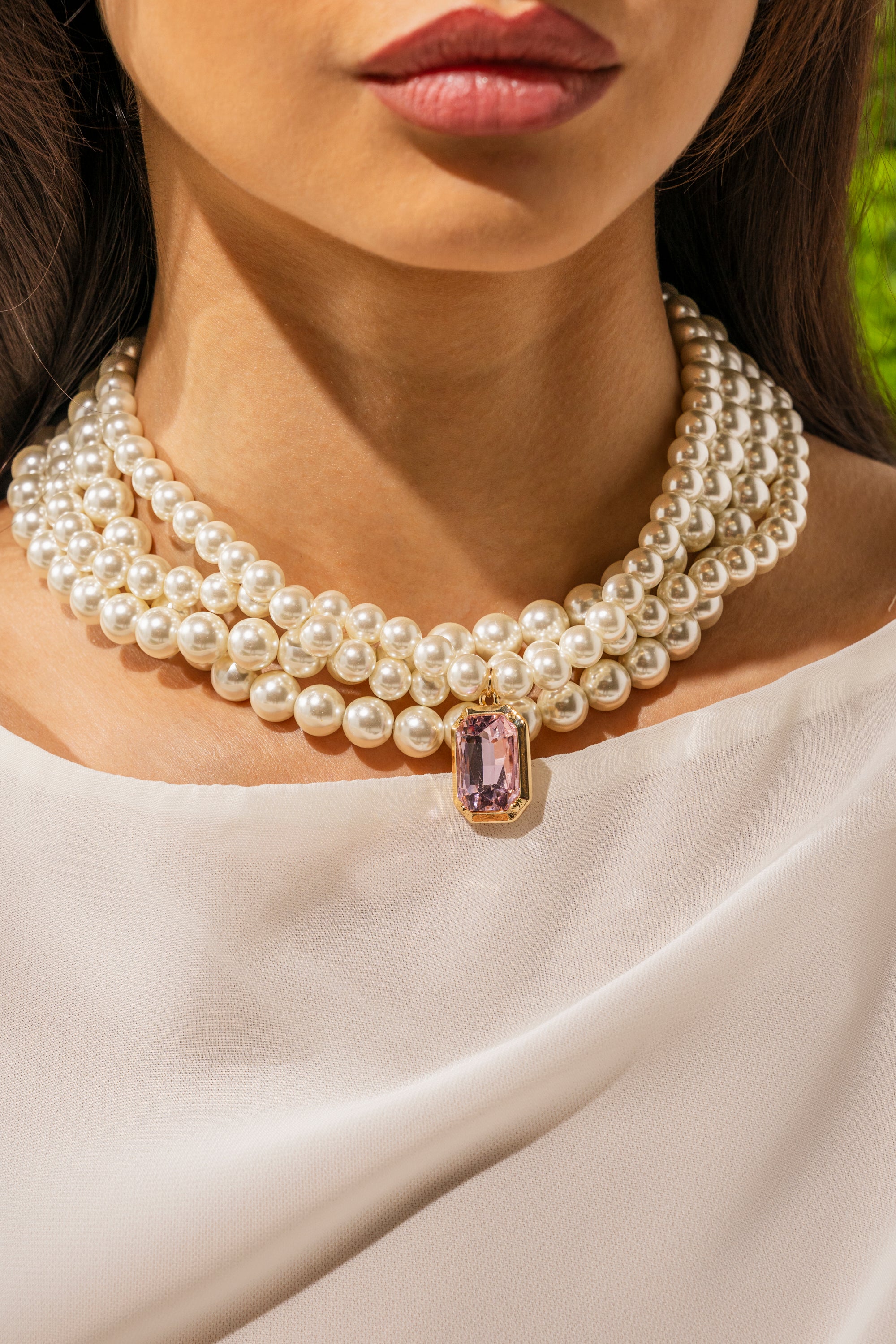 A woman wears the MARIE LAYERED NECKLACE, featuring pearls and a large rectangular pink gemstone pendant, with a white top. Her dark hair frames her face in this shoulder-up shot. 0