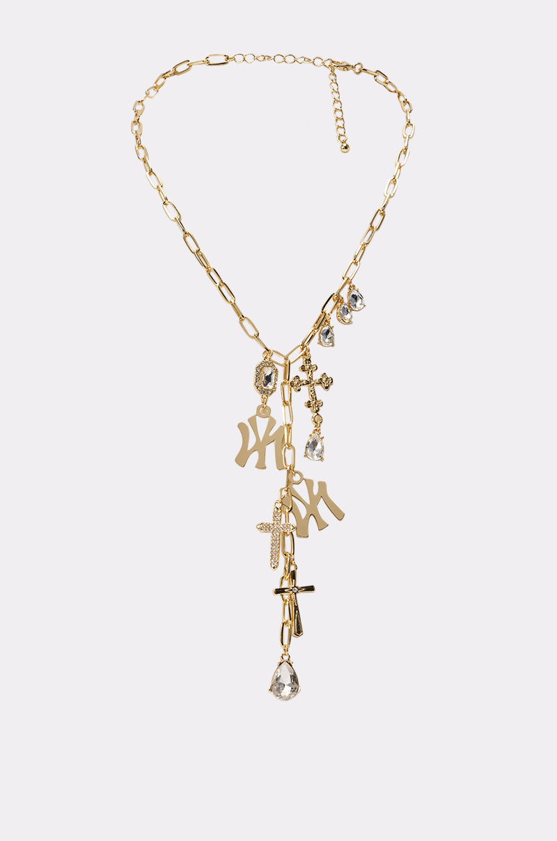 The IN YA CITY NECKLACE features gold charms, dangling letters, rhinestone crosses, a safety pin, and teardrop crystals in an asymmetrical cluster for a layered shine. 0