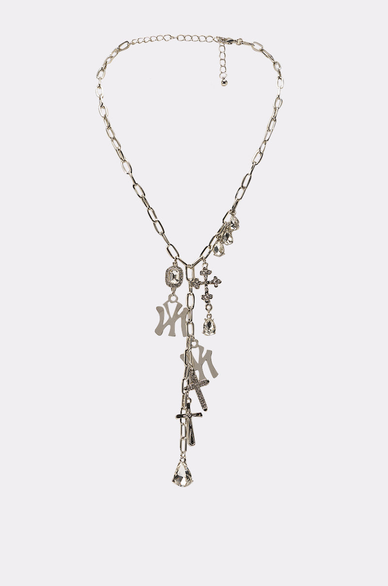 The IN YA CITY NECKLACE is a silver-tone chain-link necklace with dangling charms—letters, crystals, and geometric shapes—arranged asymmetrically. Shown against a plain, light-colored background. 0
