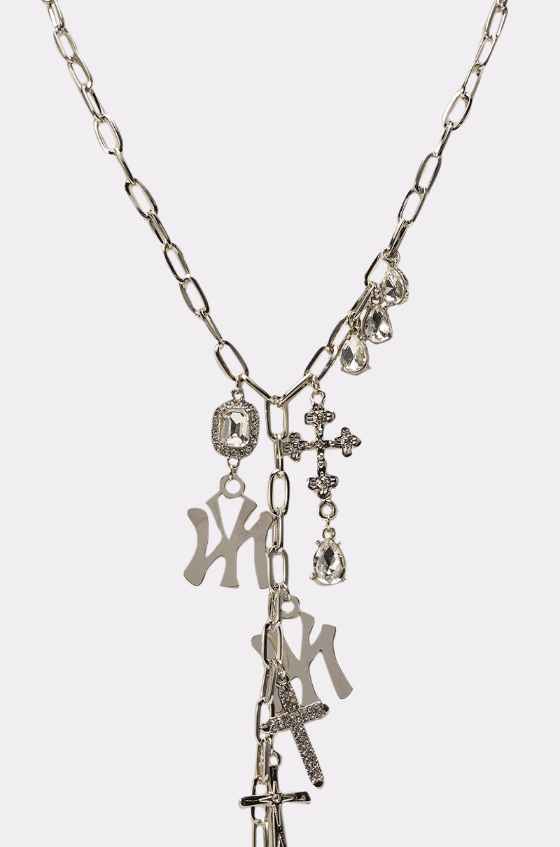 The IN YA CITY NECKLACE is a silver-tone chunky chain-link necklace with cross, crystal teardrop, and stylized letter charms, shown on a white background. 2