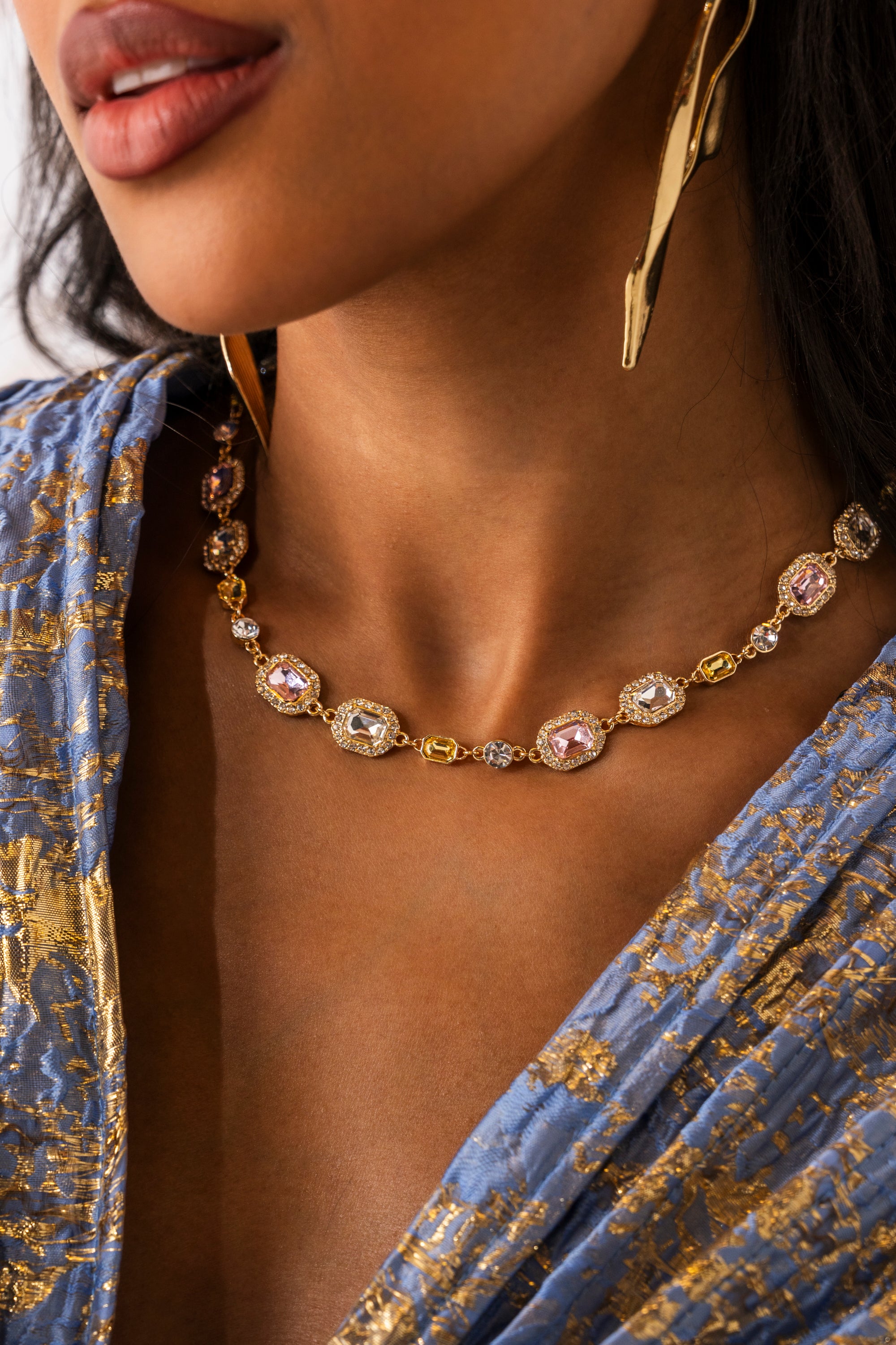 A woman wearing the WITH THE VIBES NECKLACE— a statement gold-tone piece with pink and white gemstones—plus large gold earrings and a blue-and-gold patterned garment. The focus is on her jewelry and neckline. 1