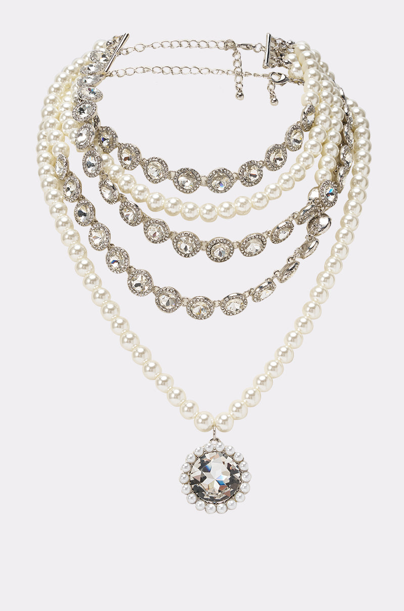 The ARI NECKLACE showcases layered strands of faux pearls and rhinestones, accented with a bold round crystal bib pendant for an elegant, formal look. 0