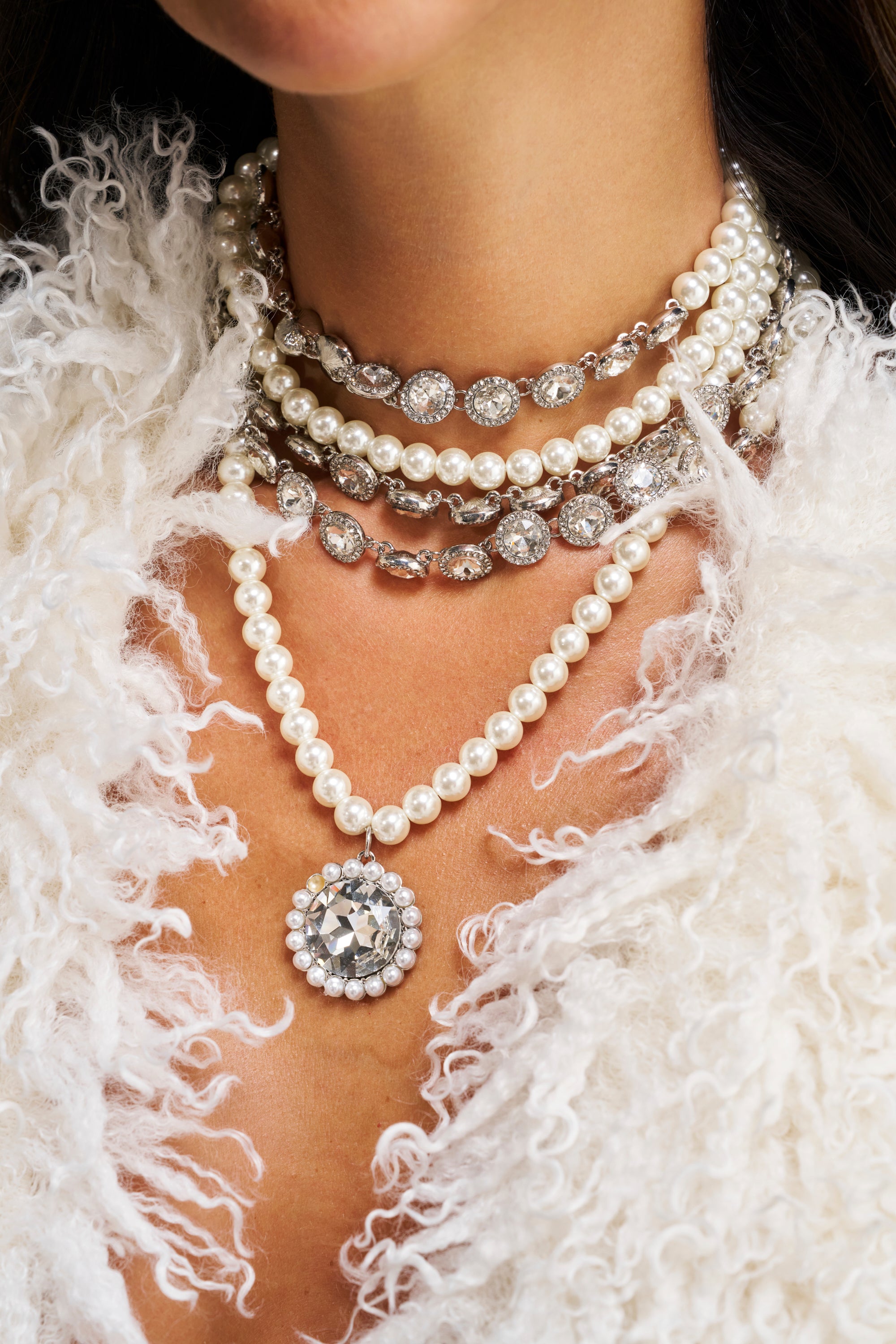 A person wears the ARI NECKLACE, a statement piece featuring pearls and jeweled details, paired with a white garment with feathery texture near the neck and chest.