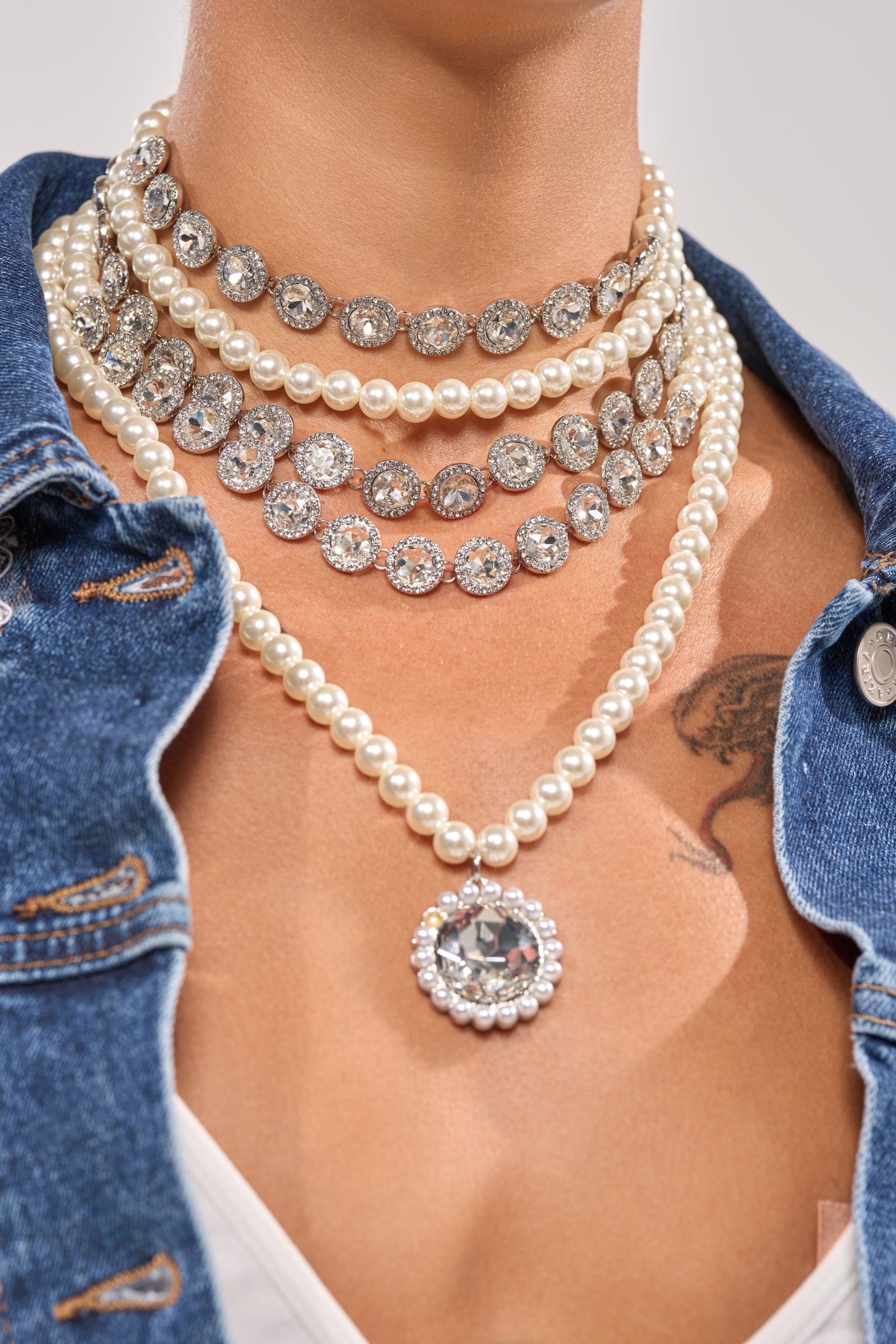 A person wears the ARI NECKLACE—a layered statement piece with pearls, rhinestones, and a large pendant—paired with a blue denim jacket, white top, and a partially visible chest tattoo. 1