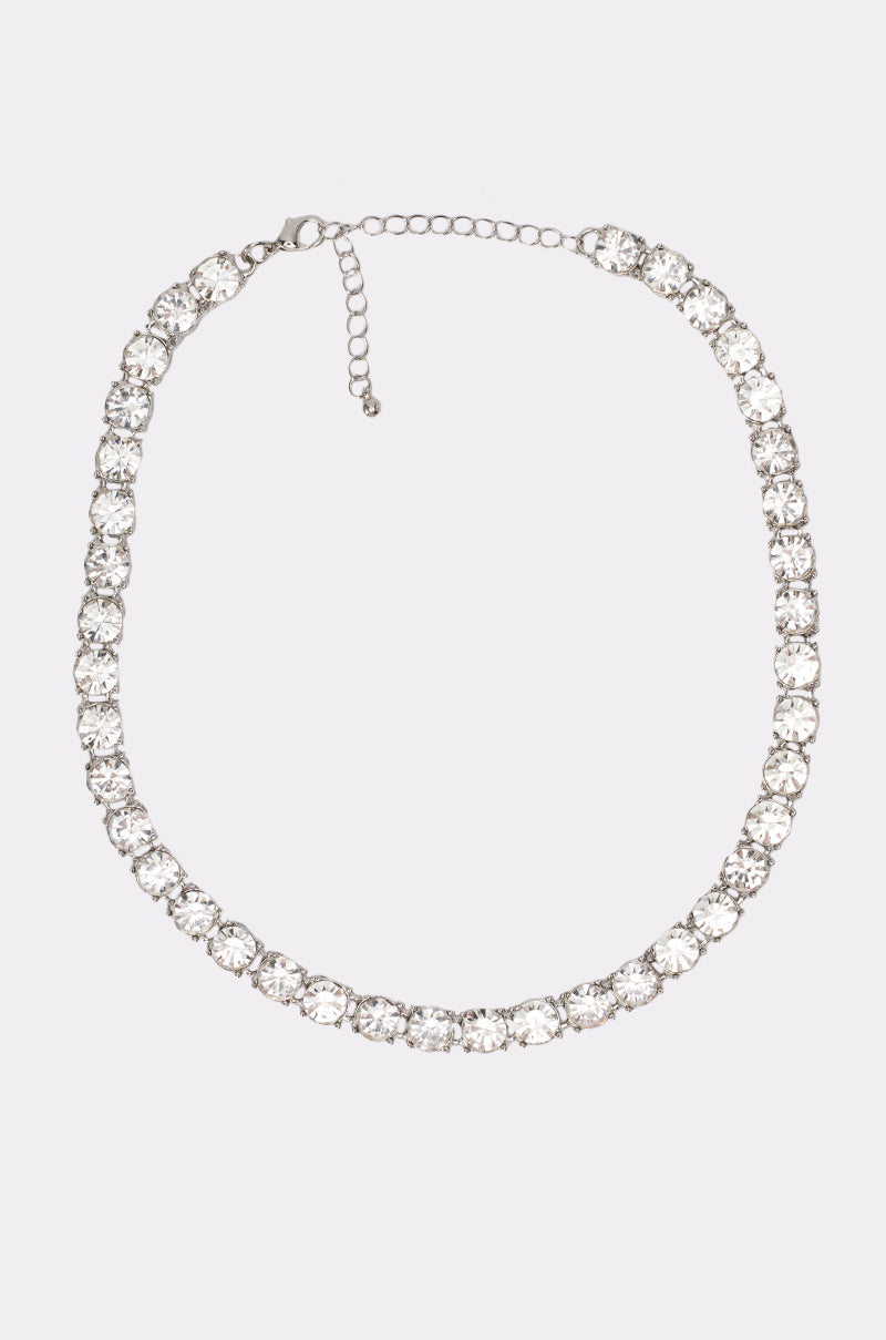 The TENNIS CHAIN NECKLACE is a silver-tone piece with a row of large, round clear rhinestones, an adjustable clasp, and is shown on a light gray background.