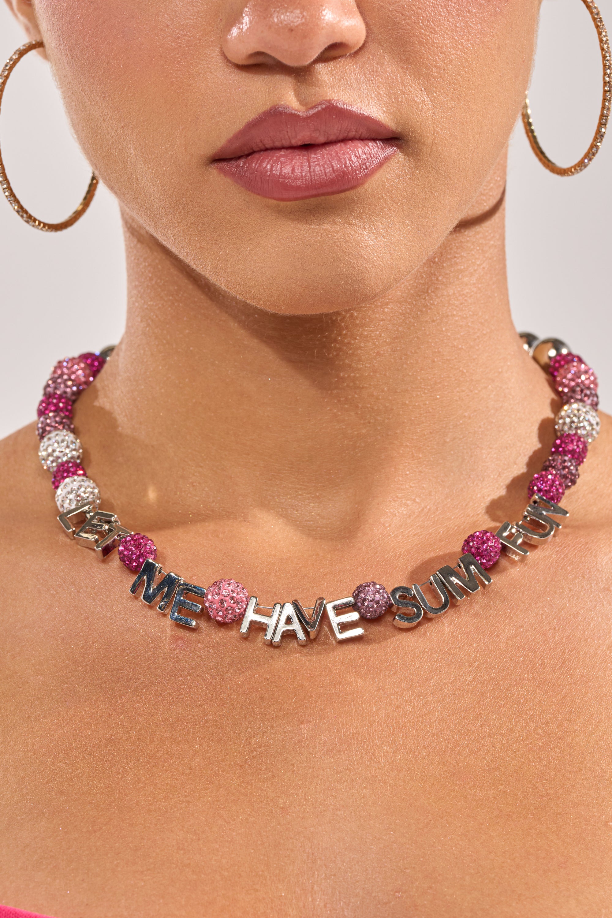A woman wears large gold hoop earrings and the WANNA HAVE FUN NECKLACE—a sparkling pink rhinestone statement piece with silver letters. The image highlights her lower face and neck. 1