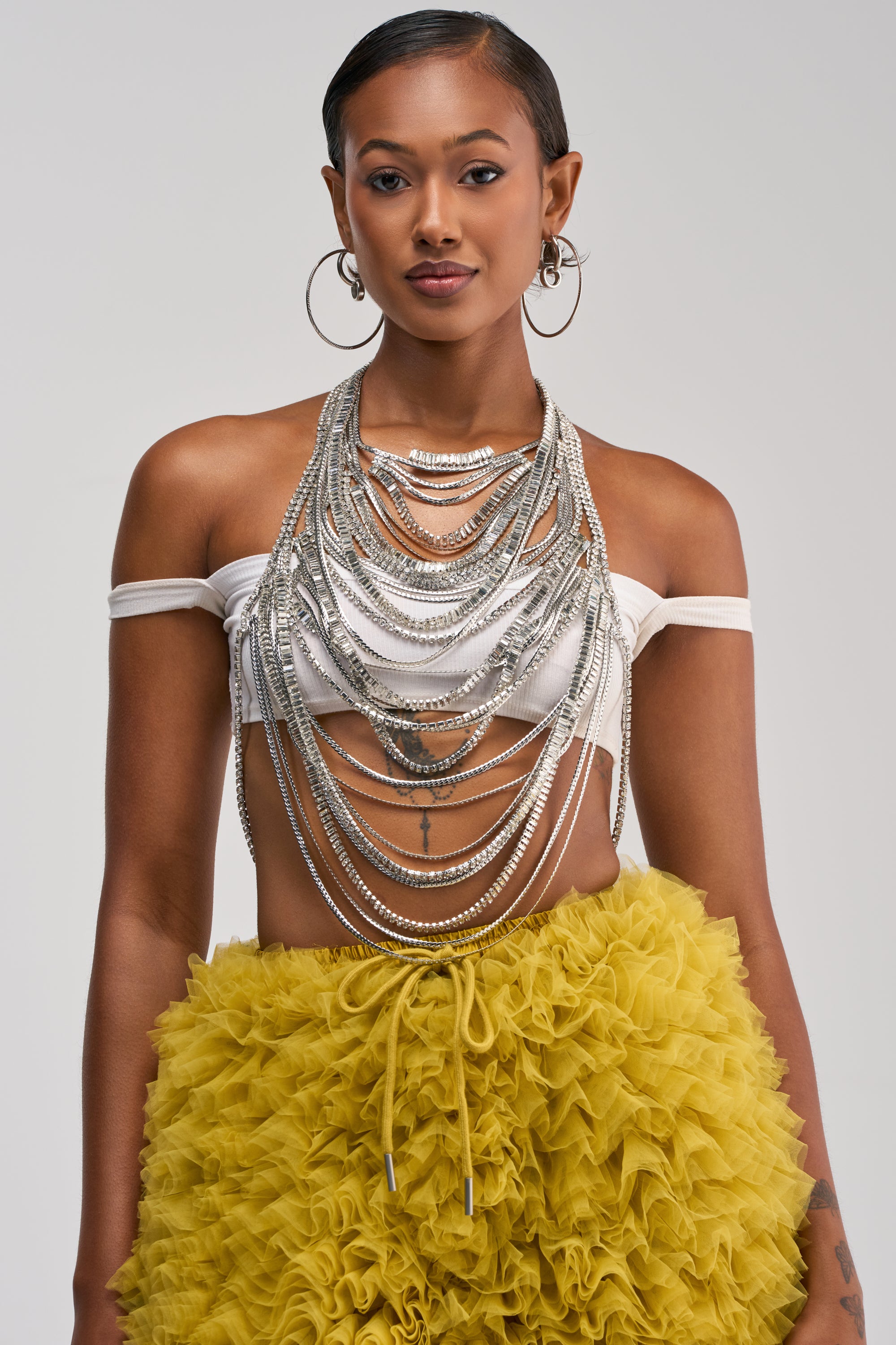Sporting a white off-shoulder crop top, large hoop earrings, and a yellow ruffled skirt, the woman completes her bold look with statement body jewelry featuring layered silver chains and the striking THAT GIRL BODYCHAIN adorned with sparkling rhinestones as she poses confidently against a plain background.